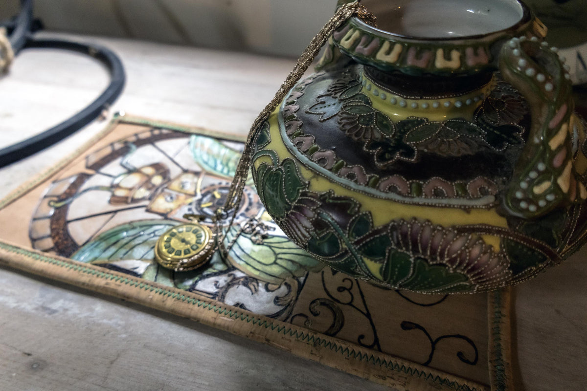 A close-up of a decorative ceramic pitcher with a gold-toned handle and lid, decorated with green, yellow, and brown floral patterns. It sits on a matching embroidered textile with a gold-toned chain and a small round gold pendant on a light wooden surface.