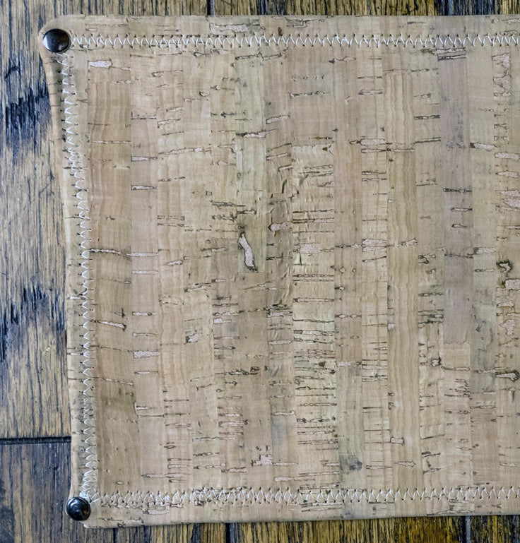 A close-up of a rectangular cork mat with stitched edges and metal corner protectors, laid flat on a dark wood surface.